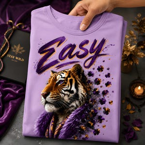 May include: A lilac-colored t-shirt featuring a tiger illustration with the word "Easy" in purple script. The tiger has orange and black stripes, purple eyes, and a purple fur collar. The shirt is folded.