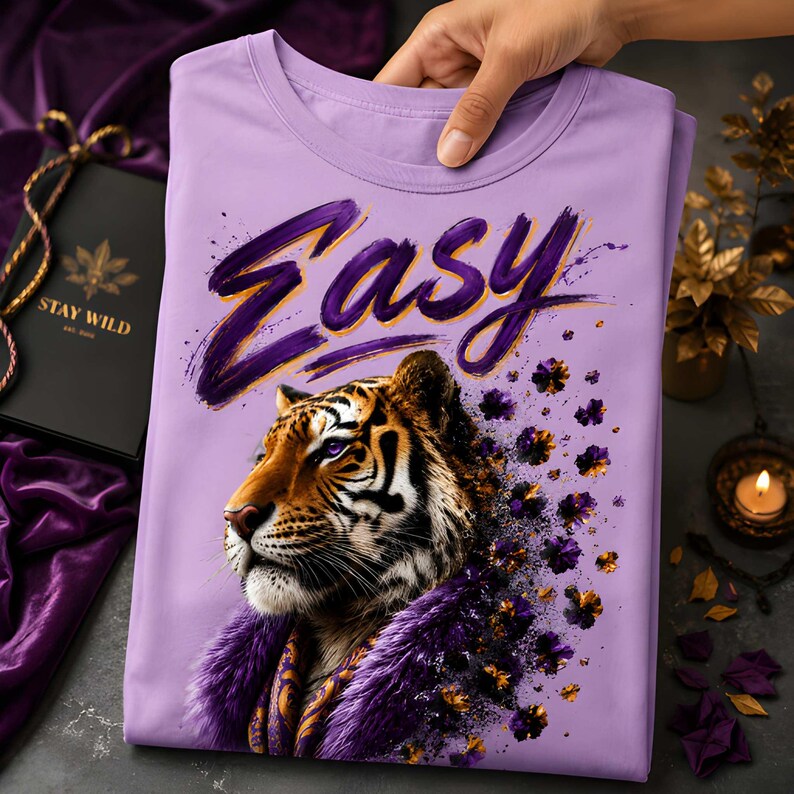 May include: A lilac-colored t-shirt featuring a tiger illustration with the word "Easy" in purple script. The tiger has orange and black stripes, purple eyes, and a purple fur collar. The shirt is folded.