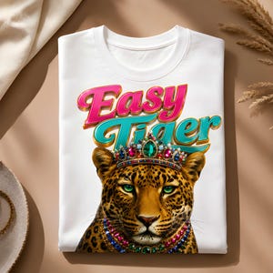 May include: White t-shirt featuring a colorful graphic of a leopard wearing a jeweled crown and necklace. The text "Easy Tiger" is written above the leopard in pink and teal lettering. The shirt is folded.