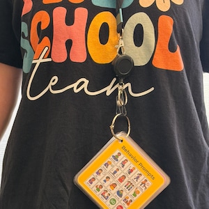 Puede incluir: Una camiseta negra con el texto PRE SCHOOL team y un gráfico de flor amarilla. Un cordón negro con un carrete retráctil sostiene una tarjeta cuadrada de plástico amarillo con una cuadrícula de iconos pequeños y el texto Behavior Prompts.