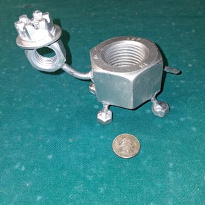 May include: A metal sculpture of a turtle made from nuts and bolts. The turtle has a large hexagonal body, a nut head, and bolt legs. A quarter is next to the turtle for size comparison.