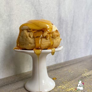 May include: A golden-brown pastry, topped with a creamy white layer and drizzled with honey, sits on a small white pedestal. Honey drips down the sides, creating a glistening effect. The dessert is presented on a wooden surface.