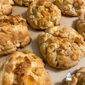 May include: Close-up of freshly baked cookies on a baking sheet. The cookies are golden brown with a crispy texture, generously topped with crunchy cornflakes. The image showcases a batch of homemade treats, perfect for dessert or a snack.