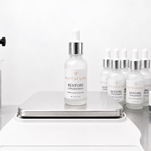 Puede incluir: Un frasco cuentagotas de vidrio esmerilado con la etiqueta Peptalume Restore Concentrate Peptide Architectural Serum y un volumen neto de 30 ml. El frasco se asienta sobre una b&aacute;scula cuadrada de acero inoxidable, rodeado de frascos id&eacute;nticos.