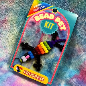 May include: A Pride Edition Bead Pet Kit featuring a rainbow colored lizard with black beads for the legs and body. The kit includes all the beads needed to make the lizard and a keychain ring. The packaging says "Bead Pet Kit" and "Progress".