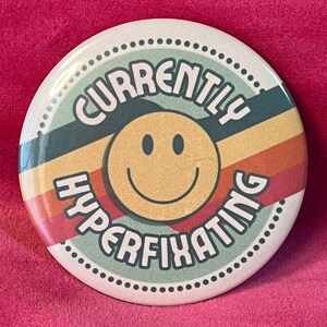 May include: A round button with a retro design featuring a smiling yellow face and the text "Currently Hyperfixating" in a green, black, and orange color scheme.