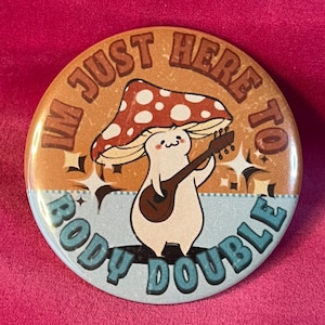 May include: A round button with a brown background and blue border. The button features a cartoon mushroom character playing a guitar with the text "I'm just here to body double" in a retro font.