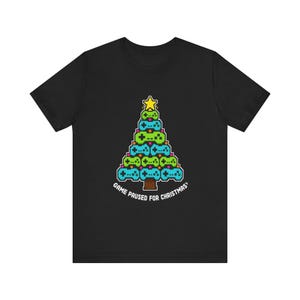 May include: A black short-sleeve t-shirt featuring a pixel art graphic of a Christmas tree constructed from green and blue video game controllers, topped with a yellow star, and the text "GAME PAUSED FOR CHRISTMAS" printed in white below.