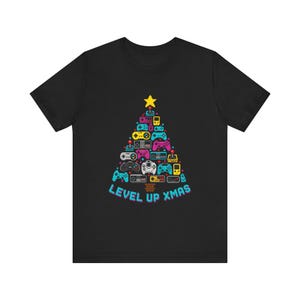 May include: A black short-sleeved t-shirt featuring a graphic of a Christmas tree constructed from various colourful pixelated video game controllers and consoles, topped with a yellow star, and the text "LEVEL UP XMAS" in blue letters.