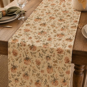 May include: A rectangular table runner featuring a cream-colored background with a repeating pattern of orange pumpkins, small brown flowers, and green leaves. The fabric has a soft, matte texture and covers the center of a wooden dining table.