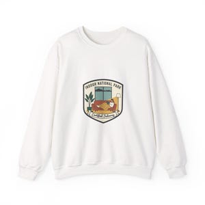 May include: White sweatshirt featuring a graphic with the words "INDOOR NATIONAL PARK" and a cosy indoor scene. The graphic includes a person on a sofa, a window, a lamp, and plants. The words "Certified Indoorsy" are also included.