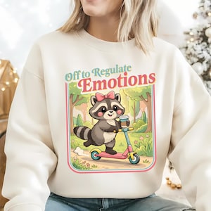 Op de afbeelding: Crèmekleurige sweatshirt met een cartoon wasbeer op een roze en blauwe scooter, met een koffiekopje. De wasbeer draagt een roze strik. De tekst "Off to Regulate Emotions" staat boven de wasbeer in een kleurrijk kader.