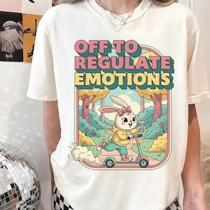 May include: Cream-colored t-shirt featuring a cartoon illustration. The graphic shows a bunny riding a scooter through a park, holding a coffee cup. Text above reads "OFF TO REGULATE EMOTIONS" in pink and yellow.
