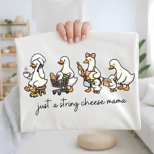 May include: Cream-coloured t-shirt with a cartoon design of four ducks engaged in different activities, such as using a phone and baking. The phrase "just a string cheese mama" is written in cursive script below the ducks.