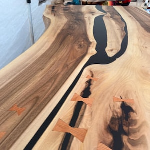 Puede incluir: Un tablero de mesa de madera con borde natural y una incrustación central de resina negra. La veta de la madera, en tonos marrones claros y oscuros, está acentuada por varias incrustaciones de madera clara en forma de mariposa en la superficie.