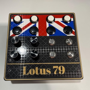 Pode incluir: Uma caixa de controle retangular com um padrão de bandeira Union Jack na metade superior e uma textura xadrez preta na inferior. Inclui quatro botões brancos, quatro pretos, quatro botões giratórios pretos e quatro botões prateados com o texto Lotus 79.