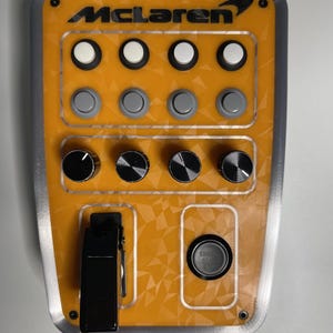 Op de afbeelding: Een McLaren knoppenpaneel met een oranje voorplaat met een geometrisch patroon. Het bevat vier witte drukknoppen, vier grijze drukknoppen, vier zwarte draaiknoppen, een zwarte tuimelschakelaar en een zwarte startknop voor de motor.