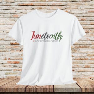 May include: White t-shirt with the word "Juneteenth" in red and green script, with the phrase "celebrate freedom" below. The design includes red and green stars. The shirt is displayed on a wooden surface.