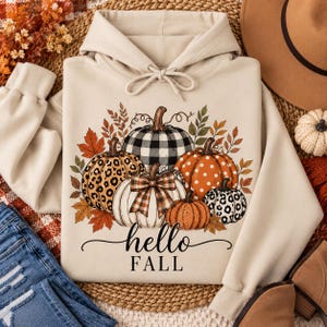 Peut inclure: Un sweat à capuche beige avec une impression graphique de cinq citrouilles aux motifs variés, incluant un carreau vichy noir et blanc, un imprimé léopard et des pois blancs, agrémenté de feuilles d'automne et du texte hello FALL.
