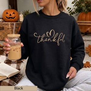 May include: A black long-sleeve crewneck sweatshirt featuring the word thankful in a gold cursive script font with a heart shape above the letter k. The garment has a ribbed neckline, cuffs, and hem, providing a simple and clean aesthetic.
