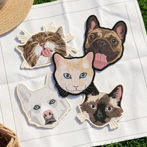 Peut inclure: Cinq écussons brodés représentant des visages d'animaux : un chat tigré brun tirant la langue, un bouledogue français beige tirant la langue, un chat crème, un chien husky blanc aux yeux bleus et un chat siamois aux marques brunes.