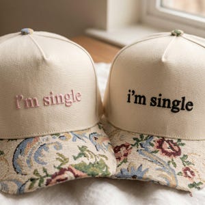 Op de afbeelding: Twee beige baseballpetten met de geborduurde tekst "I'm single". De petten hebben een bloemenpatroon in blauw, groen en rood. De tekst op één pet is roze en de andere is zwart.