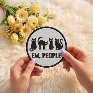 May include: A circular embroidered patch with a light gray background and a black stitched border. The design features four black cats with yellow eyes and the text "EW, PEOPLE" in bold black capital letters centered at the bottom.