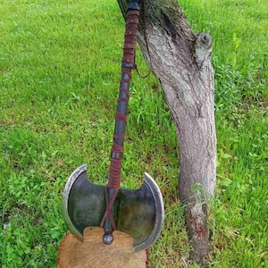 Könnte beinhalten: Eine zweischneidige Axt mit dunklem Metallkopf und einem Holzgriff, der mit braunem Leder umwickelt ist. Die Axt liegt auf einem Holzstumpf an einem Baum, mit grünem Gras im Hintergrund.