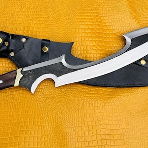 Può includere: Un coltello grande e curvo con una lama argentata lucida e un manico scuro e strutturato. Il coltello poggia su un fodero in pelle nera, con una cintura in pelle abbinata. Il manico ha dettagli dorati. Lo sfondo è una superficie gialla strutturata.