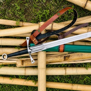 Può includere: Una spada in stile medievale con una lama argentata e un'impugnatura nera, appoggiata su steli di bambù. La spada è accompagnata da tre cinture in pelle marrone e nera, con cuciture verdi. Il fodero è verde con dettagli in pelle marrone.