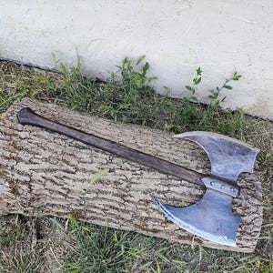 May include: A large, double-bladed axe with a dark wooden handle and a silver-colored head. The axe rests on a textured, brown log, with grass and a white wall in the background. The axe head has a curved design.