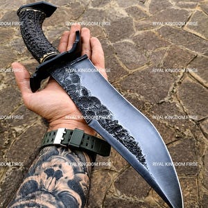 May include: A fixed-blade knife with a dark, textured, hammered steel blade and a curved, brown, antler-style handle. The knife features a black metal guard and a pommel cap. The text RIYAL KINGDOM FIRGE is repeated across the image surface.