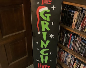 Puede incluir: Un letrero de madera negro, alto y rectangular, con las palabras "The Grinch Lives Here" en letras rojas y verdes. El letrero presenta un sombrero del Grinch y copos de nieve, con pies verdes del Grinch en la parte inferior. Una decoración festiva.