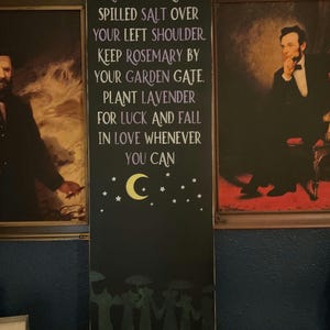 May include: A tall, rectangular black wooden sign featuring white and purple text that reads: ALWAYS THROW SPILLED SALT OVER YOUR LEFT SHOULDER, KEEP ROSEMARY BY YOUR GARDEN GATE, PLANT LAVENDER FOR LUCK AND FALL IN LOVE WHENEVER YOU CAN. Includes a yellow crescent moon, stars, and a silhouette of figures at the bottom with the text PRACTICAL MAGIC.