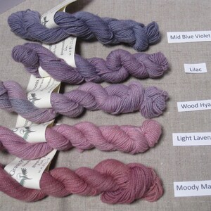 Puede incluir: Cinco madejas de hilo en tonos morados y rosas, etiquetadas como Mid Blue Violet, Lilac, Wood Hyacinth, Light Lavender y Moody Mauve. El hilo est&aacute; dispuesto sobre una superficie de lino neutra.