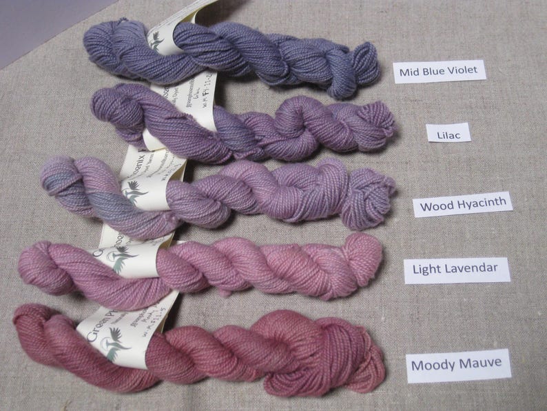 Puede incluir: Cinco madejas de hilo en tonos morados y rosas, etiquetadas como Mid Blue Violet, Lilac, Wood Hyacinth, Light Lavender y Moody Mauve. El hilo est&aacute; dispuesto sobre una superficie de lino neutra.