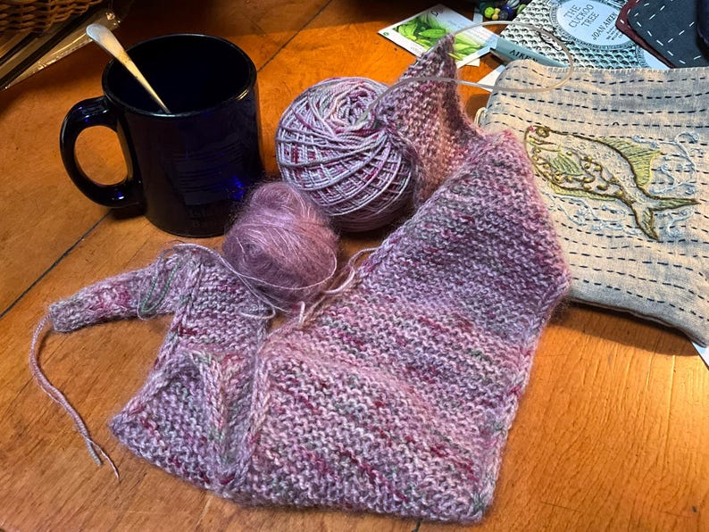 Puede incluir: Una taza azul oscuro con una cuchara, ovillos de lana y una prenda parcialmente tejida en tonos de rosa, morado y verde. Tambi&eacute;n hay una peque&ntilde;a bolsa bordada con un dise&ntilde;o de pez.