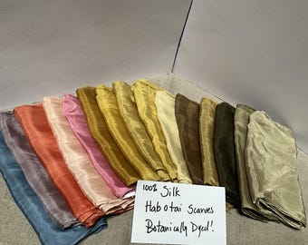 Puede incluir: Una colección de pañuelos de seda Habotai plegados en varios colores, incluyendo azul, morado, naranja, rosa, amarillo, dorado, marrón y verde. Una tarjeta blanca en primer plano presenta el texto escrito a mano: 100% Silk Habotai Scarves Botanically Dyed!.