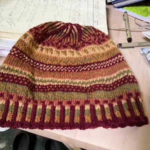 Puede incluir: Un gorro tejido en tonos burdeos, dorado y verde. El gorro presenta un patr&oacute;n de rayas con intrincados detalles de tejido. El borde es de un burdeos m&aacute;s oscuro y la corona tiene un dise&ntilde;o texturizado. Este gorro hecho a mano es perfecto para el invierno.