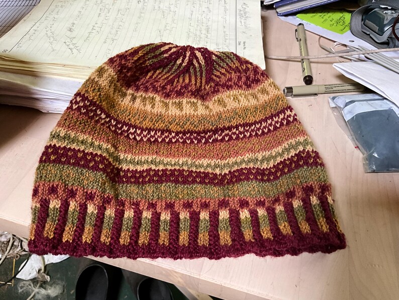 Puede incluir: Un gorro tejido en tonos burdeos, dorado y verde. El gorro presenta un patr&oacute;n de rayas con intrincados detalles de tejido. El borde es de un burdeos m&aacute;s oscuro y la corona tiene un dise&ntilde;o texturizado. Este gorro hecho a mano es perfecto para el invierno.