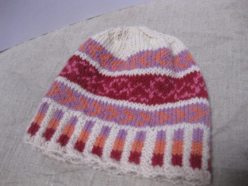 Puede incluir: Un gorro de punto crema, con bandas de patrones geom&eacute;tricos rojos, rosas, naranjas y morados. El gorro presenta un borde acanalado y un aspecto suave, adecuado para el clima fr&iacute;o.