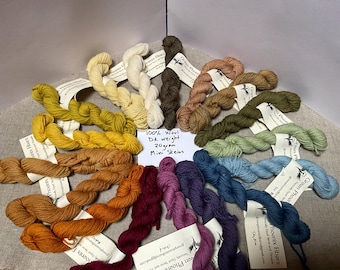 Puede incluir: Una colección de mini madejas de lana 100% DK de 20g dispuestas en círculo. Cada madeja tiene una etiqueta de papel blanco con el texto Green Phoenix Fibers. Los colores del hilo incluyen amarillo, crema, marrón, beige, verde, azul y morado.