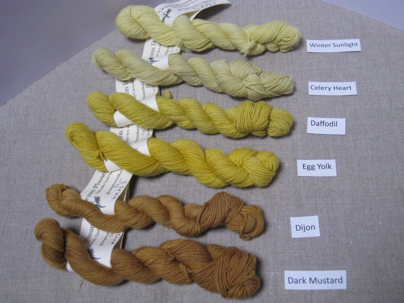 Puede incluir: Cinco madejas de hilo te&ntilde;ido a mano en tonos amarillos y marrones. Los colores est&aacute;n etiquetados como "Winter Sunlight", "Celery Heart", "Daffodil", "Egg Yolk", "Dijon" y "Dark Mustard". El hilo se muestra sobre una superficie de color neutro.