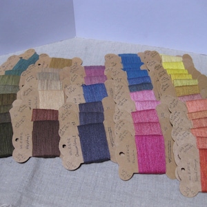 May include: A collection of colorful thread samples arranged in a fan shape. The threads are attached to small, scalloped cardboard tags with handwritten labels. The color palette includes shades of green, blue, pink, yellow, and orange.
