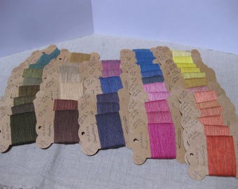 Puede incluir: Una colección de muestras de hilo de colores dispuestas en forma de abanico. Los hilos están unidos a pequeñas etiquetas de cartón festoneadas con etiquetas manuscritas. La paleta de colores incluye tonos de verde, azul, rosa, amarillo y naranja.