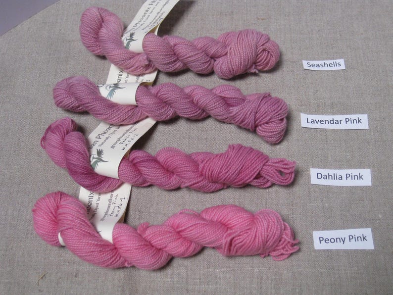 Puede incluir: Cuatro madejas de hilo rosa en diferentes tonos, etiquetadas como Seashells, Lavendar Pink, Dahlia Pink y Peony Pink. Cada madeja est&aacute; torcida y tiene una etiqueta. El hilo se muestra sobre una superficie neutra y texturizada.