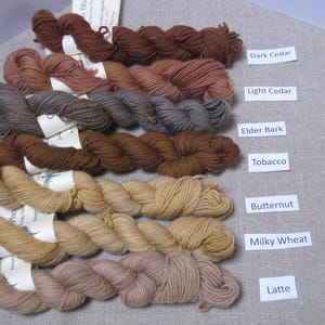 Puede incluir: Una colecci&oacute;n de madejas de hilo en tonos marrones, beige y tostados, etiquetadas con nombres como "Dark Cedar" y "Latte". El hilo est&aacute; dispuesto sobre una superficie de color neutro, mostrando la variedad de colores de fibra natural.