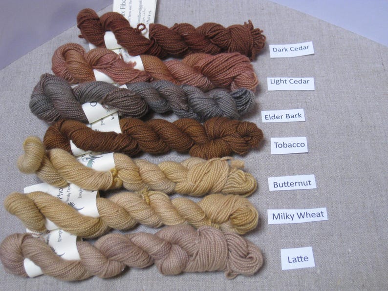 Puede incluir: Una colecci&oacute;n de madejas de hilo en tonos marrones, beige y tostados, etiquetadas con nombres como "Dark Cedar" y "Latte". El hilo est&aacute; dispuesto sobre una superficie de color neutro, mostrando la variedad de colores de fibra natural.