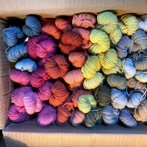 Puede incluir: Una caja de cart&oacute;n llena de madejas de hilo de colores. El hilo est&aacute; dispuesto en una variedad de colores, incluyendo azules, rosas, naranjas, verdes y grises. Las madejas est&aacute;n bien enrolladas y parecen suaves, lo que sugiere que est&aacute;n hechas de lana o una fibra similar.