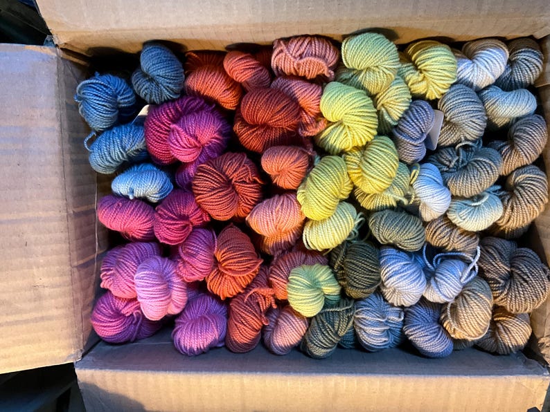 Puede incluir: Una caja de cart&oacute;n llena de madejas de hilo de colores. El hilo est&aacute; dispuesto en una variedad de colores, incluyendo azules, rosas, naranjas, verdes y grises. Las madejas est&aacute;n bien enrolladas y parecen suaves, lo que sugiere que est&aacute;n hechas de lana o una fibra similar.